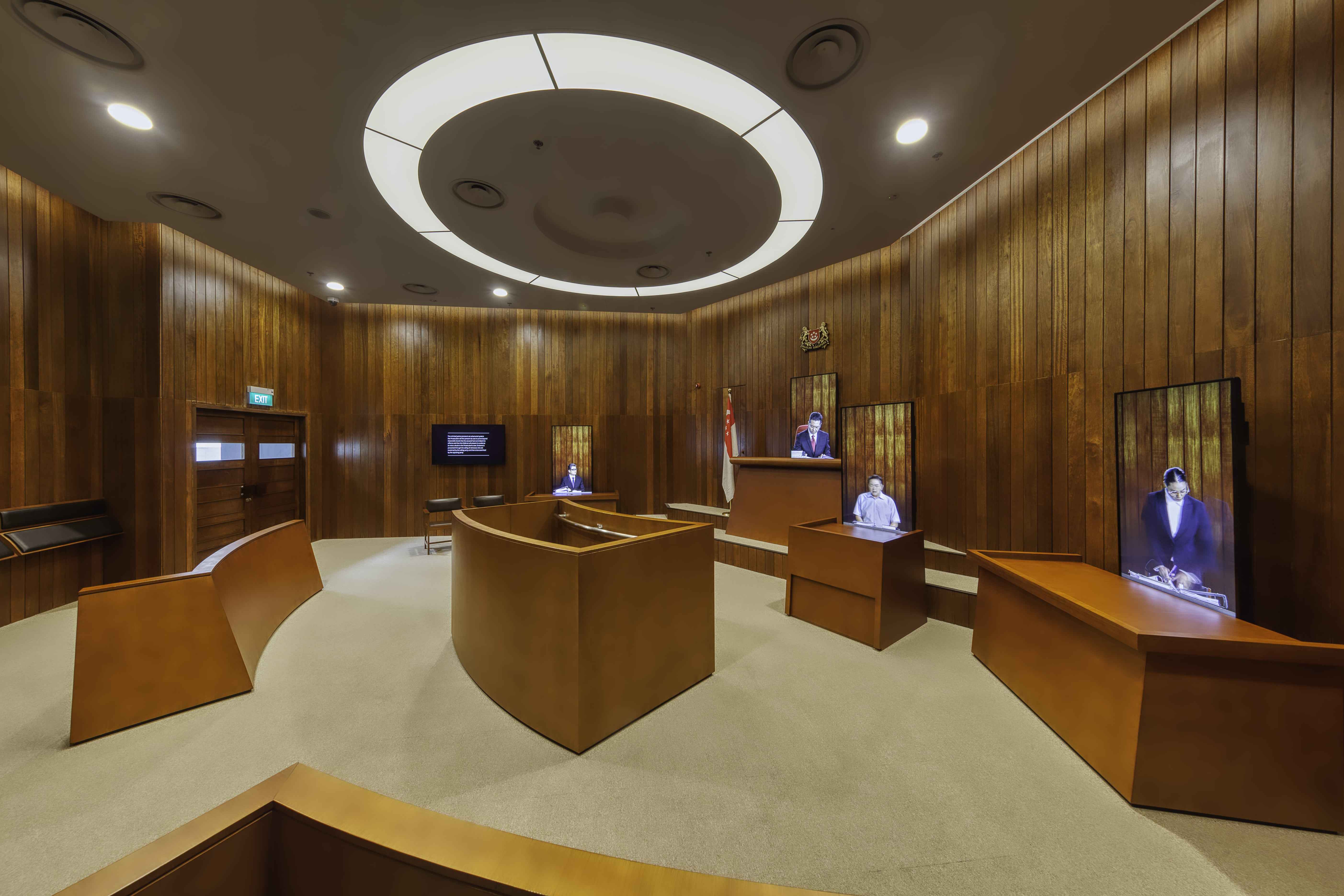 A wood-paneled courtroom with curved desks, a circular ceiling light, and screens displaying people in legal proceedings.